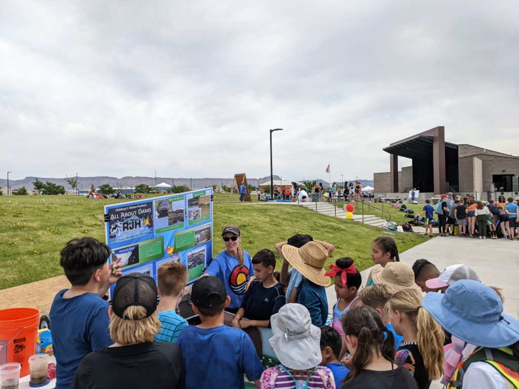 A group of children gathers around a display board outdoors, listening to an adult speak. The event takes place on a grassy area near a building, with other groups and activities visible in the background under a cloudy sky.