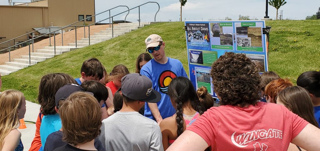 A group of students stands outside, gathered around a man in a blue shirt and cap who is presenting information on a display board about dams and rivers. The scene is sunny with grass and buildings in the background.