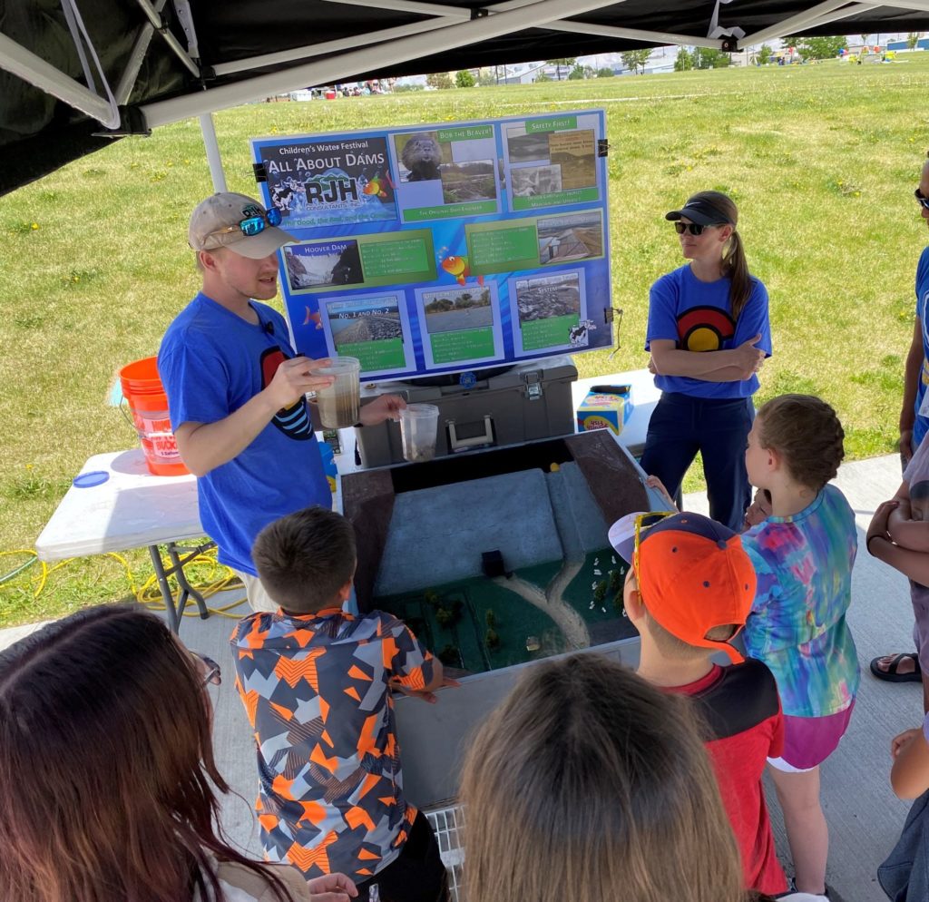 A group of children gathers around a man demonstrating a dam model at an outdoor educational booth, while another adult looks on. Informational posters about dams are displayed behind them on a sunny day.