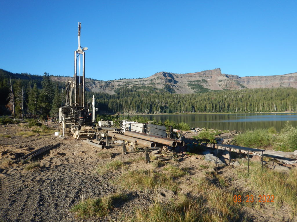 A drilling rig and equipment are set up on the shore of a lake, surrounded by grassy terrain and pine trees, with forested mountains and clear blue sky in the background. The date stamp reads 08-23-2022.