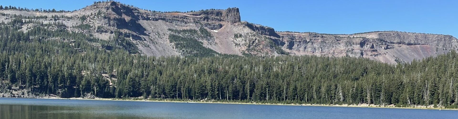 Clear blue lake with rocky shoreline in the foreground, surrounded by dense forest and distant flat-topped mountains under a cloudless blue sky. The mountain and trees are reflected in the calm water.