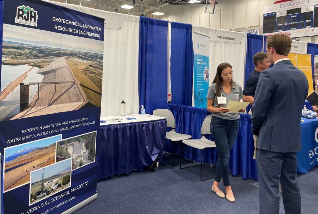 A woman holding papers stands near a trade show booth with a “Geotechnical and Water Resources Engineering” banner. She speaks with a man in a suit; other booths and people are visible in the background.