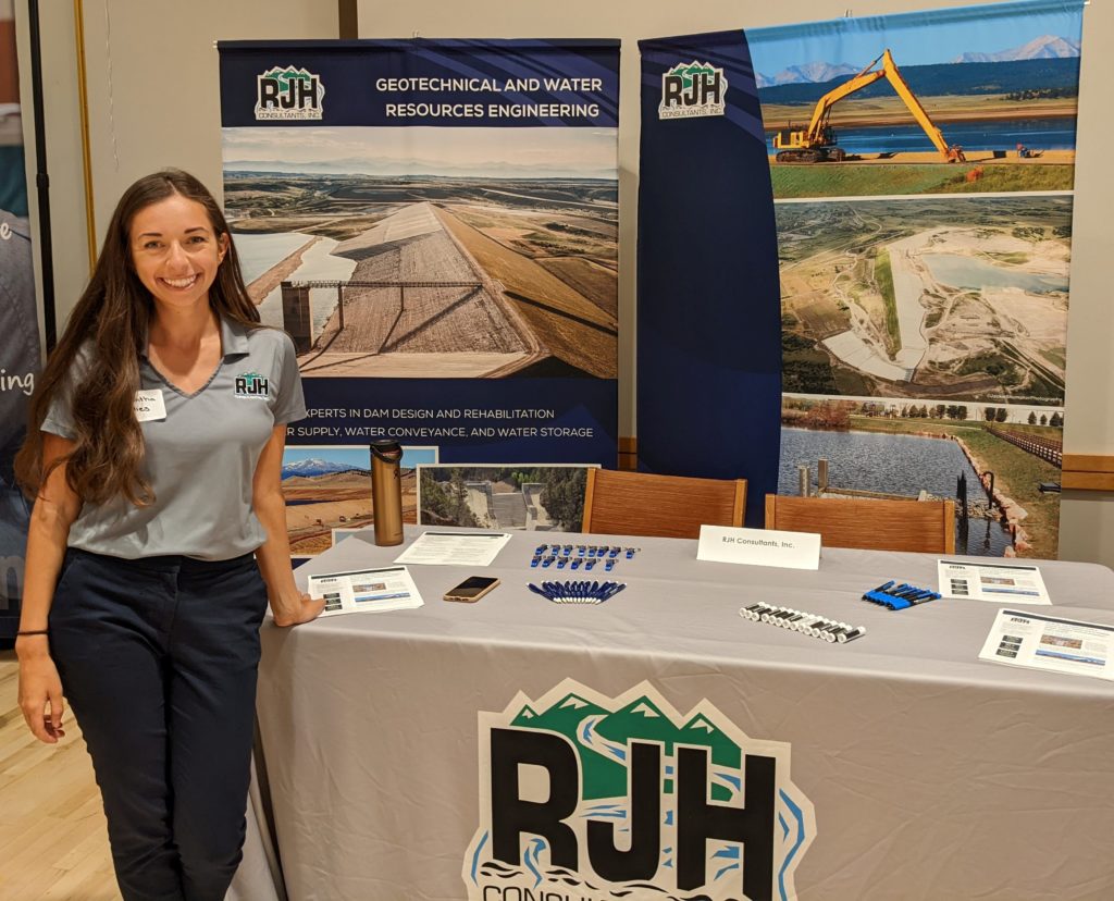 A woman stands smiling next to a booth for RJH Consultants, featuring brochures, pens, and display boards about geotechnical and water resources engineering, with large photos of infrastructure projects in the background.