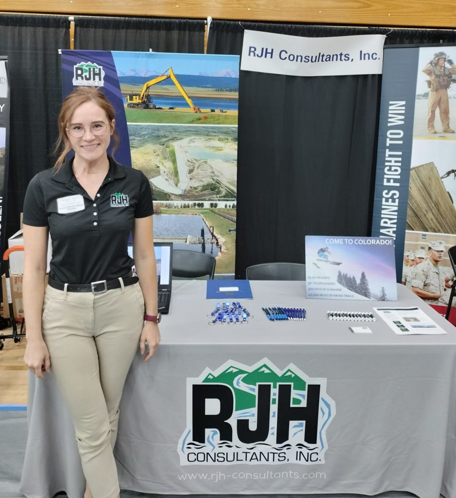 A woman in business casual attire stands and smiles in front of an RJH Consultants, Inc. booth with informational materials, brochures, and a laptop on the table at an indoor event.