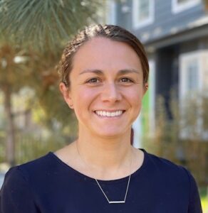 A woman with brown hair pulled back smiles at the camera. She is wearing a navy blue top and a simple necklace. The background shows trees and a blurred building on a sunny day.