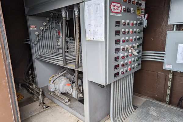 Industrial control panel with numerous buttons, switches, and indicator lights next to a complex arrangement of pipes, valves, and a small motor. A schematic is posted on the panel; the floor is concrete with some exposed wiring.